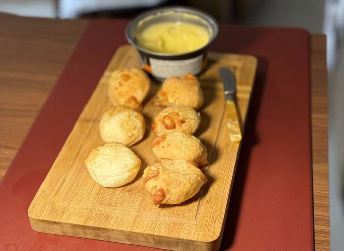 7 pães de queijo tamanho P, servidos em uma tábua, com um pote de manteiga parcialmente desfocado ao fundo