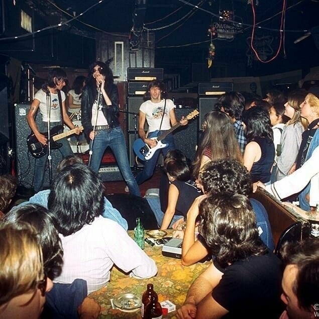 Cena interna de casa noturna. Em uma mesa diretamente em frente ao palco, vários homens e mulheres, de costas para a câmera, assistem ao show de uma banda com 4 integrantes, cujo vocalista está de jaqueta preta e óculos escuros, e os demais músicos estão de camisetas brancas, todos de jeans.