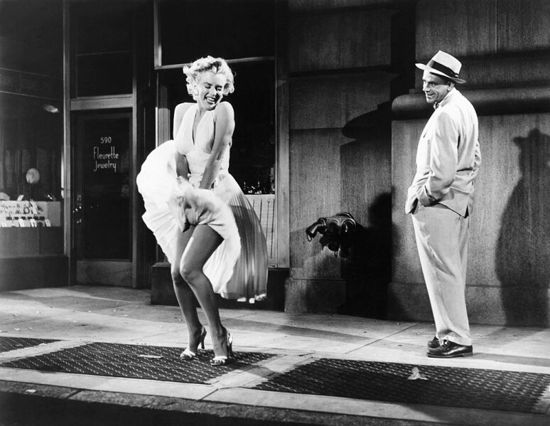 Marilyn Monroe, no estúdio, sorri e segura o vestido levantado pela saída de ar no chão abaixo dela