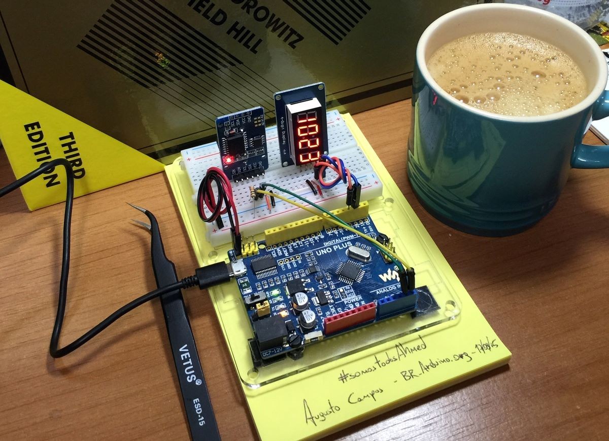 Um Arduino Uno “plus“ montado ao lado de uma mini breadboard na qual estão espetadas duas plaquinhas, sendo que uma delas tem dígitos de relógio iluminados.