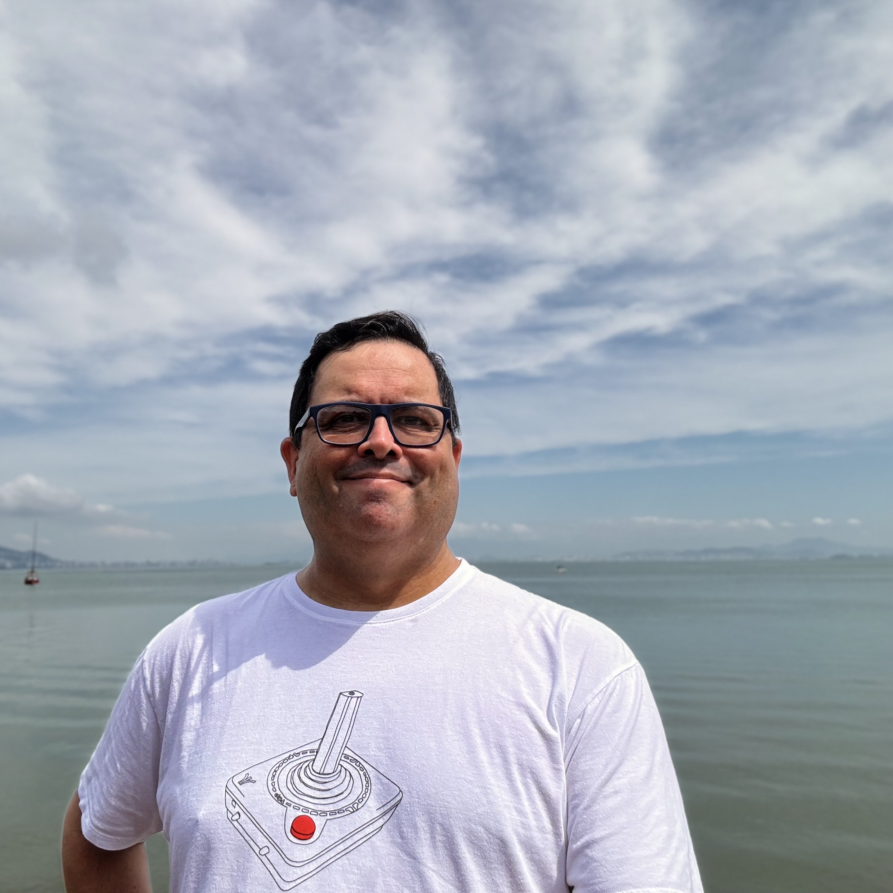 Foto minha olhando para a câmera, de camiseta estampada com um joystick de Atari, com o mar e o céu parcialmente nublado ao fundo. 