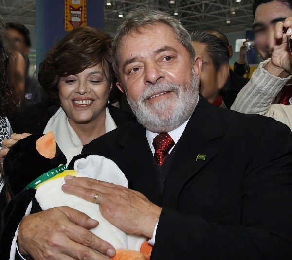 O Presidente Lula, em evento de tecnologia, segura um pinguim de pelúcia com um colar verde e amarelo com o nome de uma distribuição Linux nacional