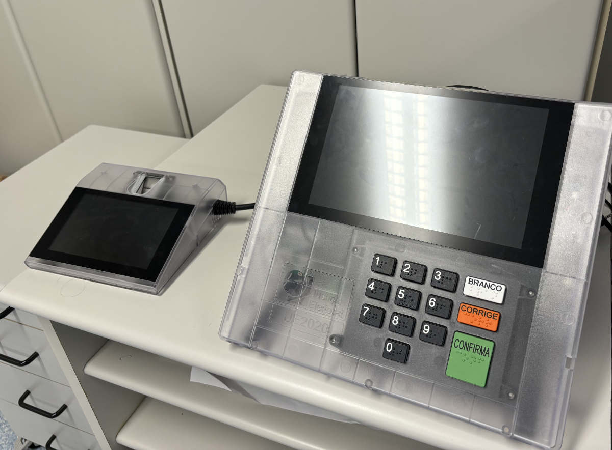 Urna eletrônica modelo 2020, com gabinete translúcido permitindo visão parcial dos componentes de seu interior, posicionada em cima de uma escrivaninha.