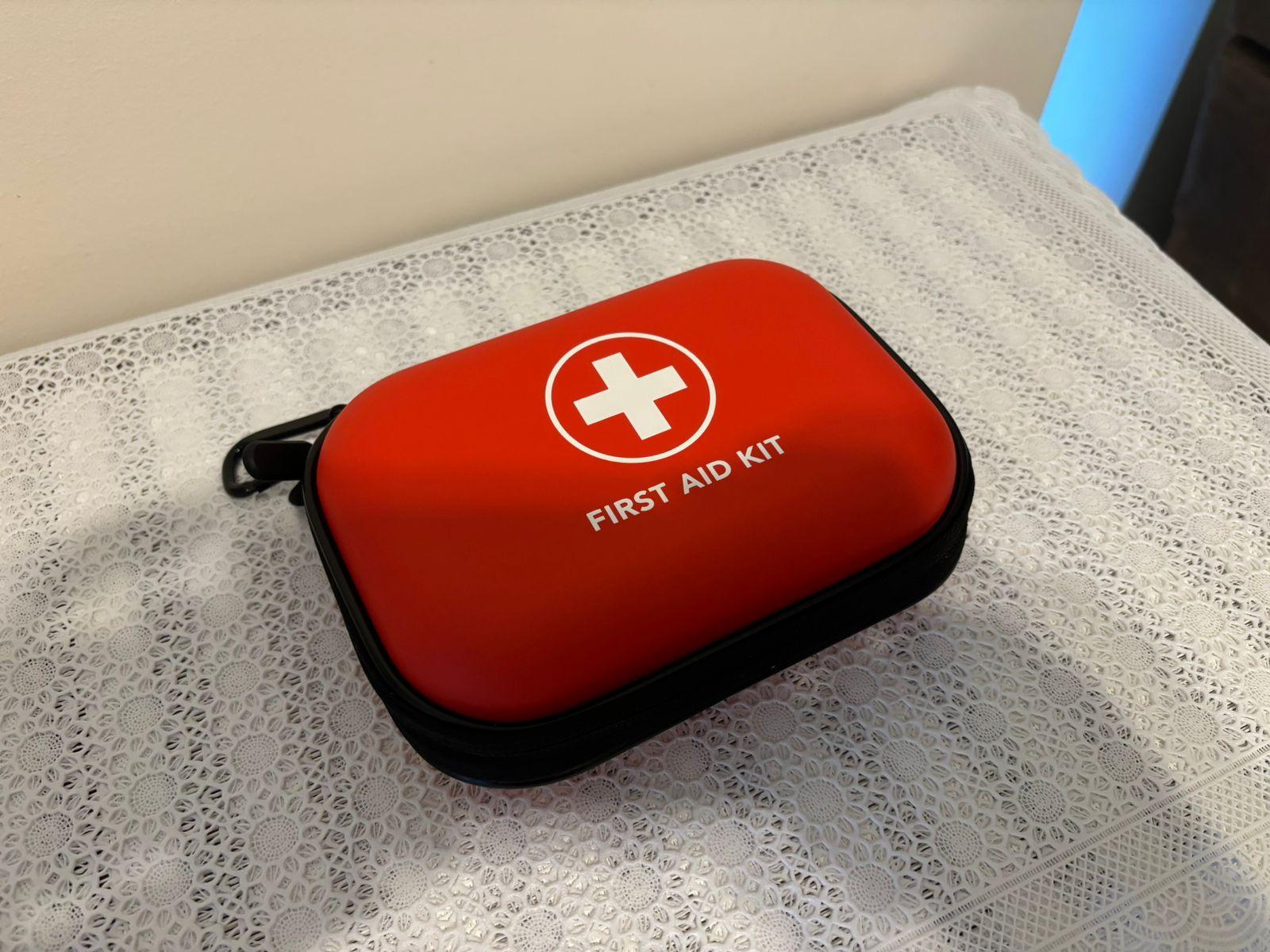 Um estojo vermelho com zíper, em formato de paralelepípedo achatado e com arestas arredondadas, onde está escrito, em letras brancas, FIRST AID KIT