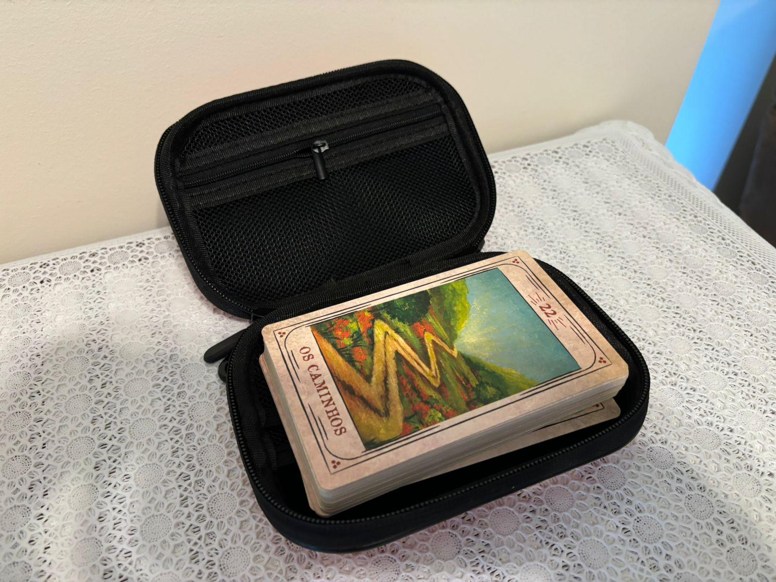Interior do mesmo estojo descrito na outra foto anexa a este post, em cor preta, com um bolso com zíper, e contendo um baralho, em cujo topo está a carta 22 - “Os Caminhos”.