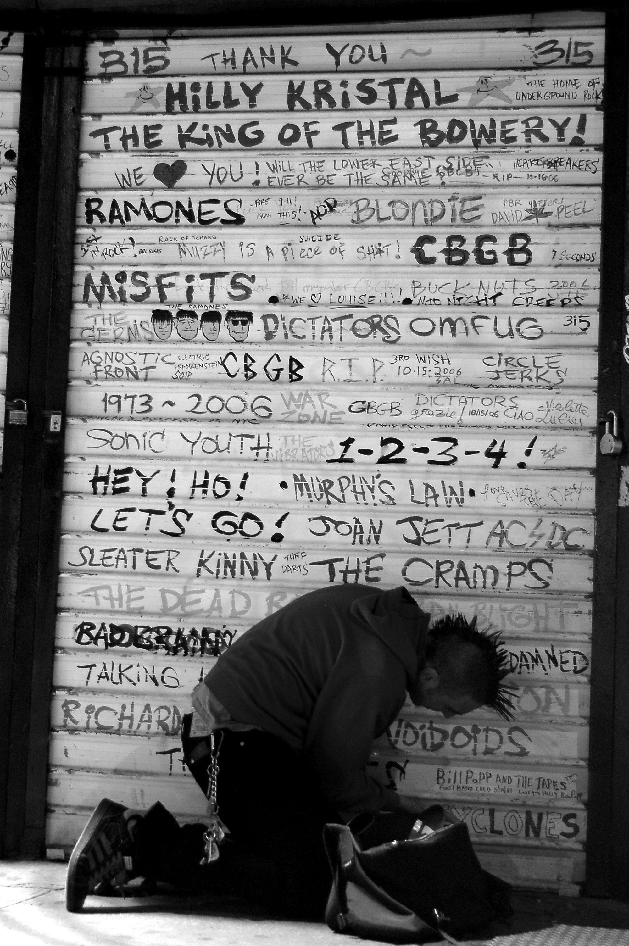 Um punk ajoelhado junto à porta fechada do CBGB, decorada com os nomes de várias das bandas e artistas que neles se apresentaram entre os anos 70 e o início do século XXI.
