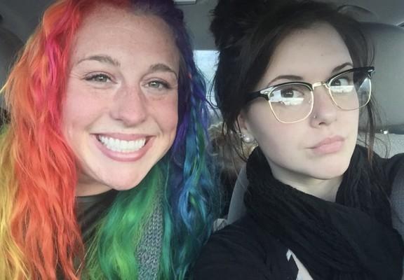 Foto dos rostos de duas irmãs dentro do carro, olhando para a câmera. Uma delas, com sorriso largo, tem os cabelos multicoloridos. A outra, com expressão contida, usa óculos e um piercing discreto no nariz, tem os cabelos pretos e está toda vestida de preto.