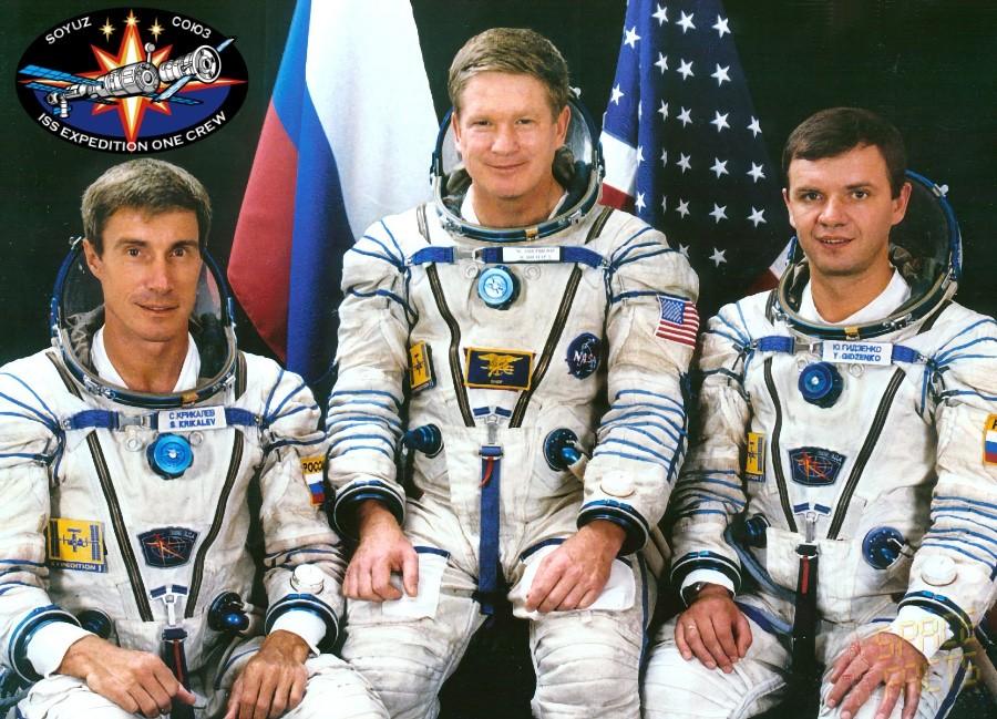 Foto oficial dos 3 integrantes trajados para o espaço, e emblema da missão Soyuz TM-31