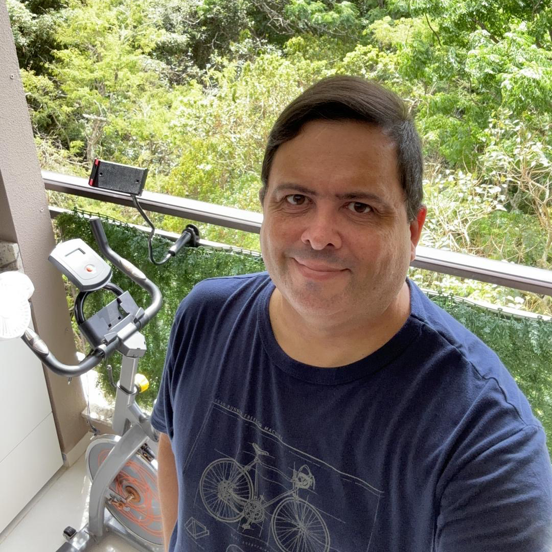 Foto minha, sorrindo par a câmera, vestindo uma camiseta azul com estampa de bicicleta, e tendo ao fundo uma bicicleta ergométrica e a mata.