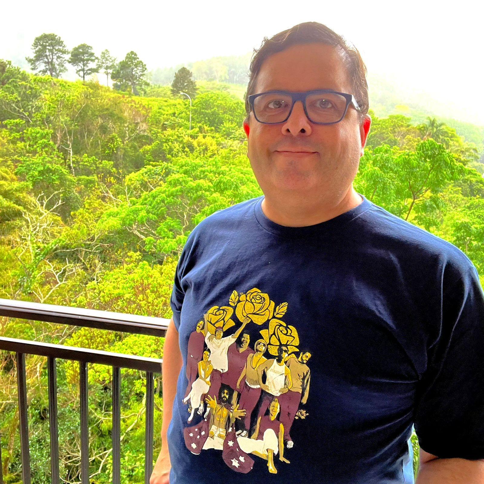 Foto minha na sacada, com a mata e a chuva ao fundo, olhando para a câmera, com camiseta azul cuja estampa mostra Raul Seixas, Tim Maia, Belchior, Cássia Eller e outros nomes brasileiros da música que já se foram