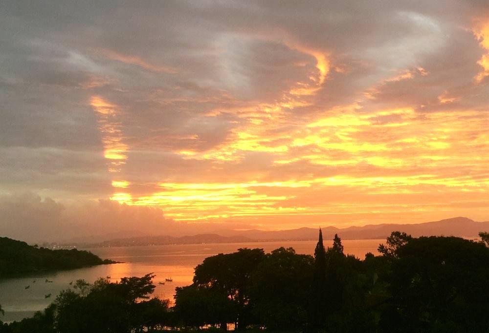 foto de um por do sol cheio de dourados e avermelhados, por cima da Baía Norte de Florianópolis, com o portinho de pescadores do bairro João Paulo visível entre árvores.