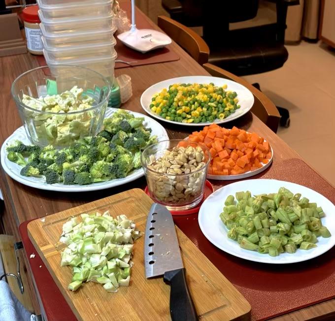 legumes cortados em uma tábua, cercada por pratos contendo vagens, brócolis, cenoura e couve-flor, todos também cortados, e mais um contendo ervilhas e milho (grãos) e outro contendo amendoins sem casca.