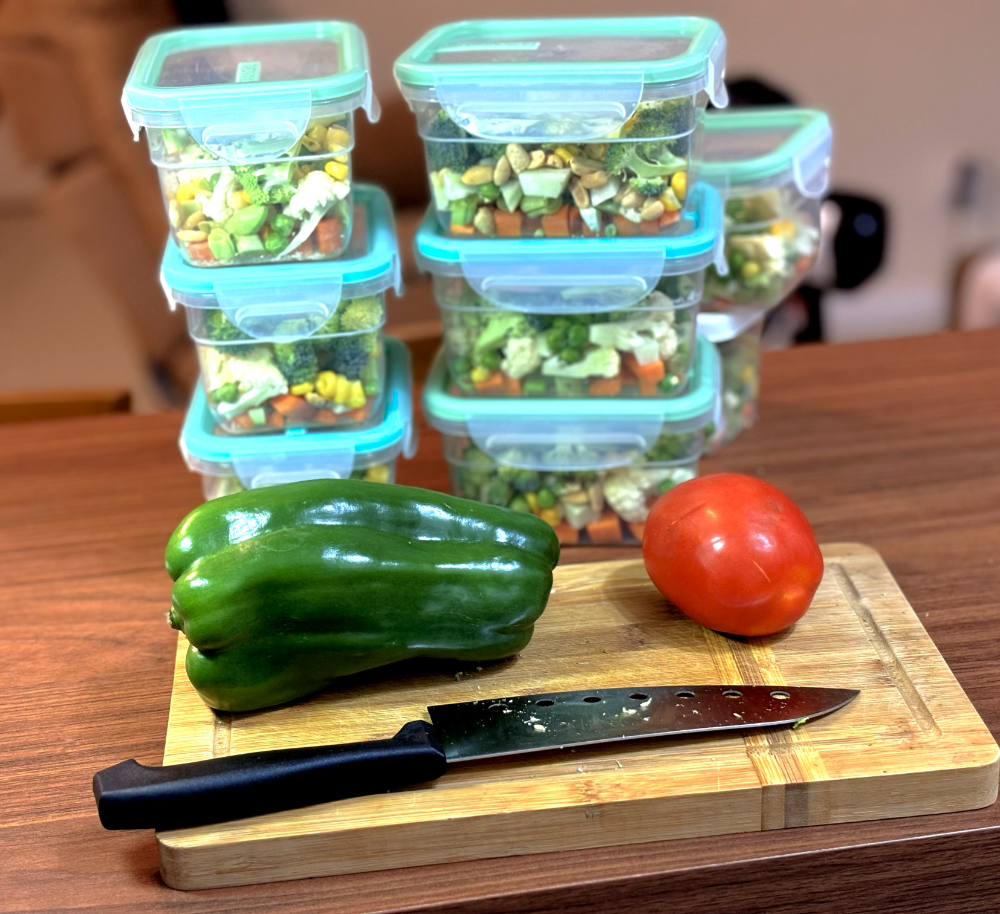 Oito potes herméticos empilhados, contendo vegetais picados em cubinhos parcialmente visíveis devido ao material translúcido, e posicionados atrás de uma tábua de corte onde estão uma faca, um pimentão verde e um tomate.