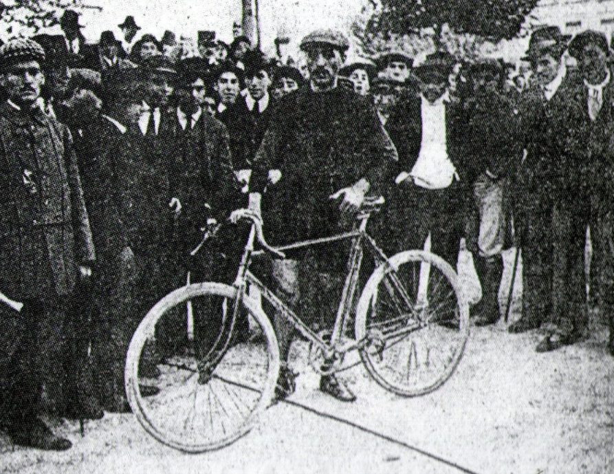 O vencedor Charles George, com sua bicicleta, rodeado por homens de chapéu ou boné, em foto em preto e branco