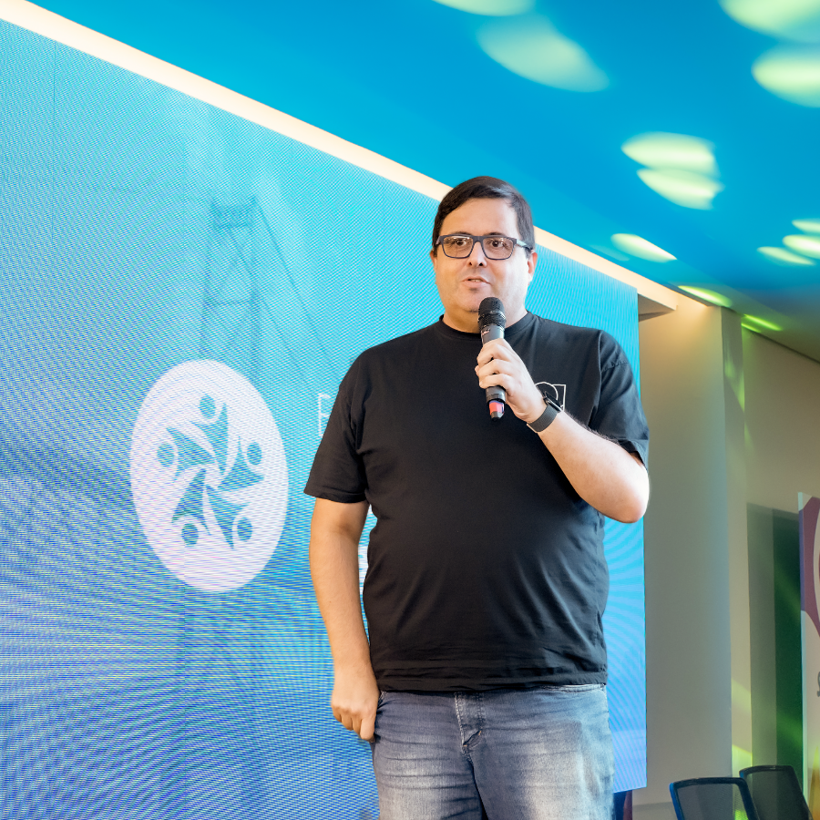 Foto minha, com um painel de apresentação ao fundo, de jeans e camiseta preta com um logo apenas parcialmente visível e que não permite a identificação, e segurando um microfone sem fio.