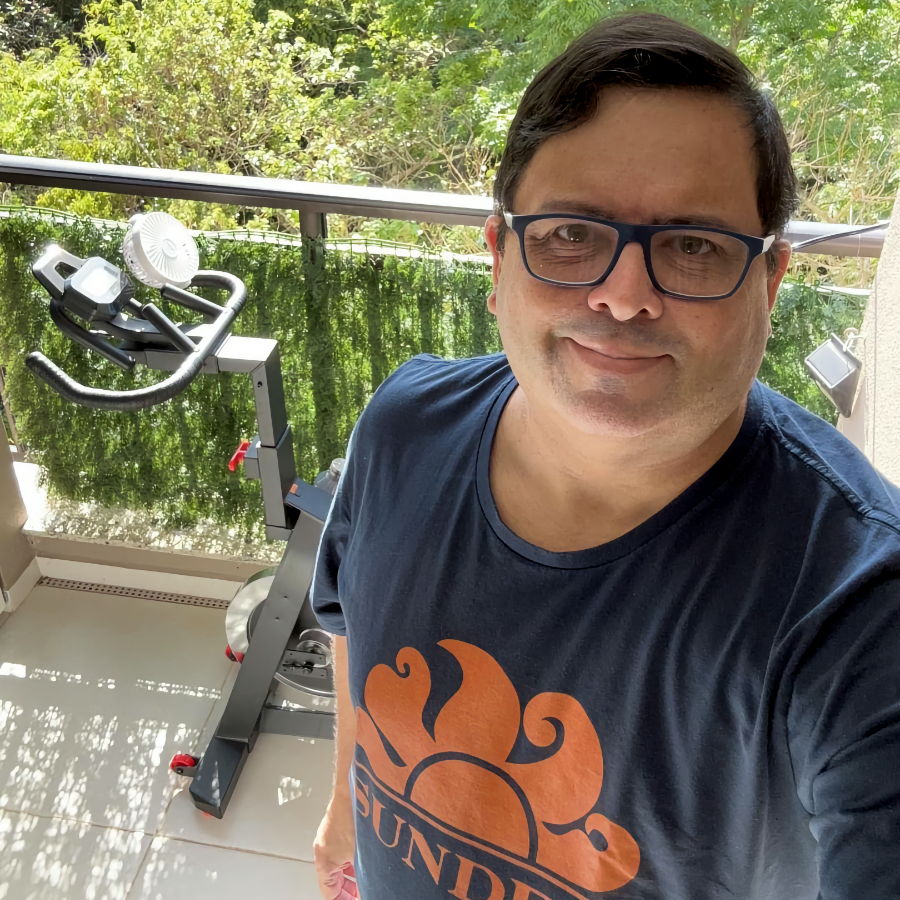 Foto minha olhando pra câmera, com camiseta azul com o logo da marca Sundek, dos anos 80. Atrás de mim está parcialmente visível uma bicicleta indoor e, ao fundo, está a mata. 