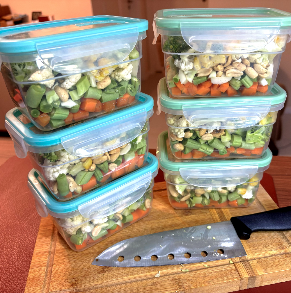 duas pilhas de 3 potes herméticos fechados, cada um deles contendo cenoura, vagem, couve-flor, brócolis, amendoim, milho e ervilha. As pilhas estão posicionadas em uma tábua de corte, na qual também está visível uma faca.