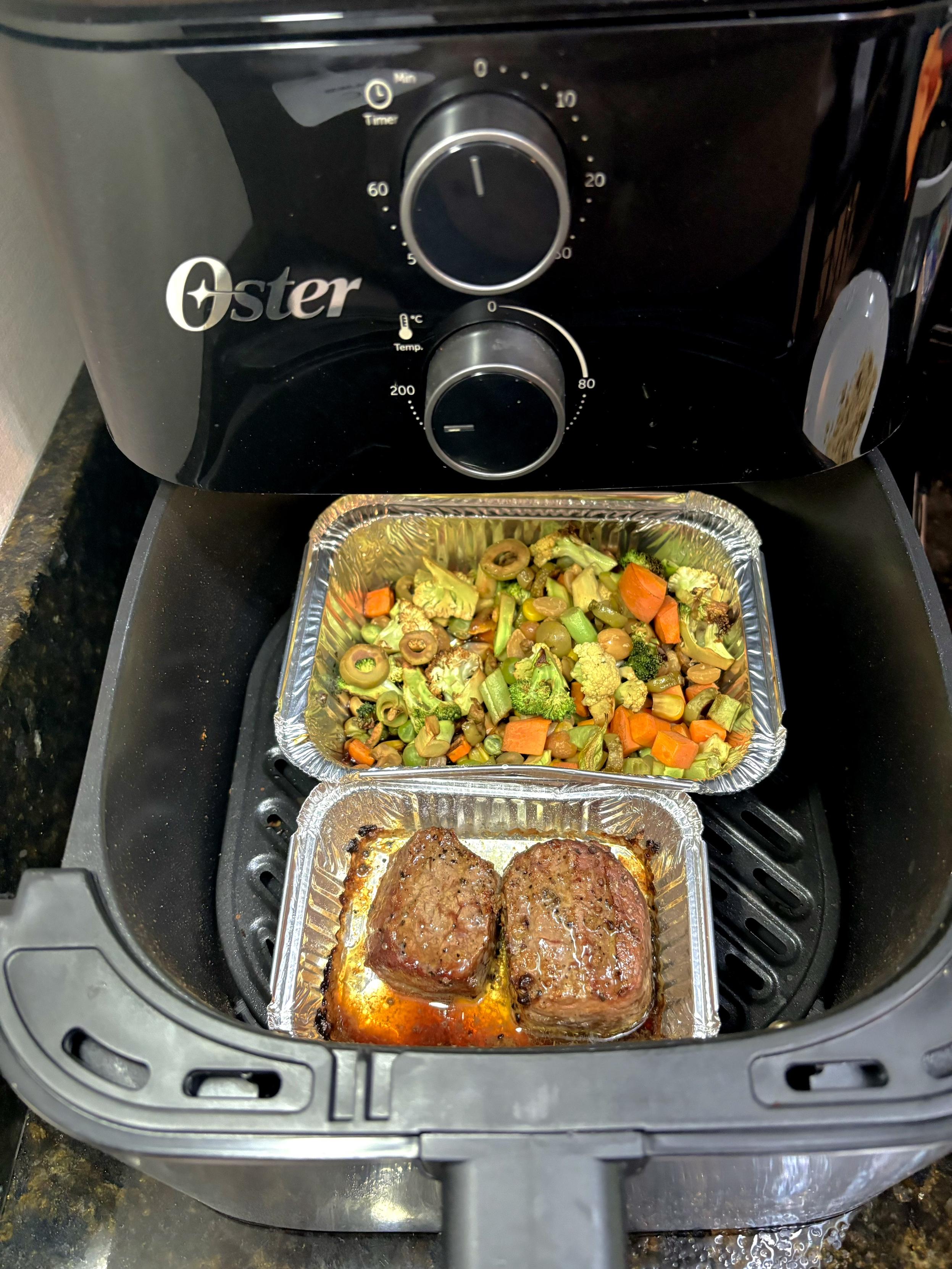 Dois recipientes de alumínio dentro de uma airfryer. O menor deles tem dois medalhões de carne bovina, e o maior tem vegetais picados. Os itens estão com cores e aparência de estarem prontos para o consumo. 