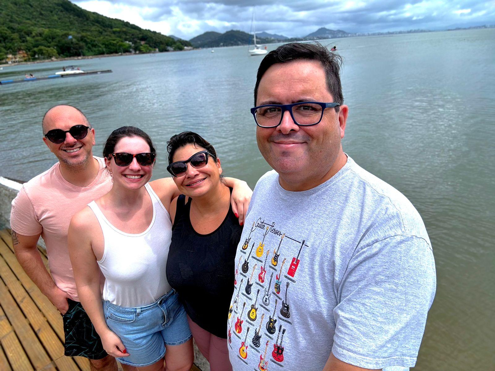 Com o mar e um veleiro ao fundo, olham para a câmera (da esquerda para a direita) um homem, duas mulheres, e um segundo homem. Todos menos o segundo homem estão de óculos escuros. O segundo homem usa uma camiseta estampada com guitarras elétricas coloridas.