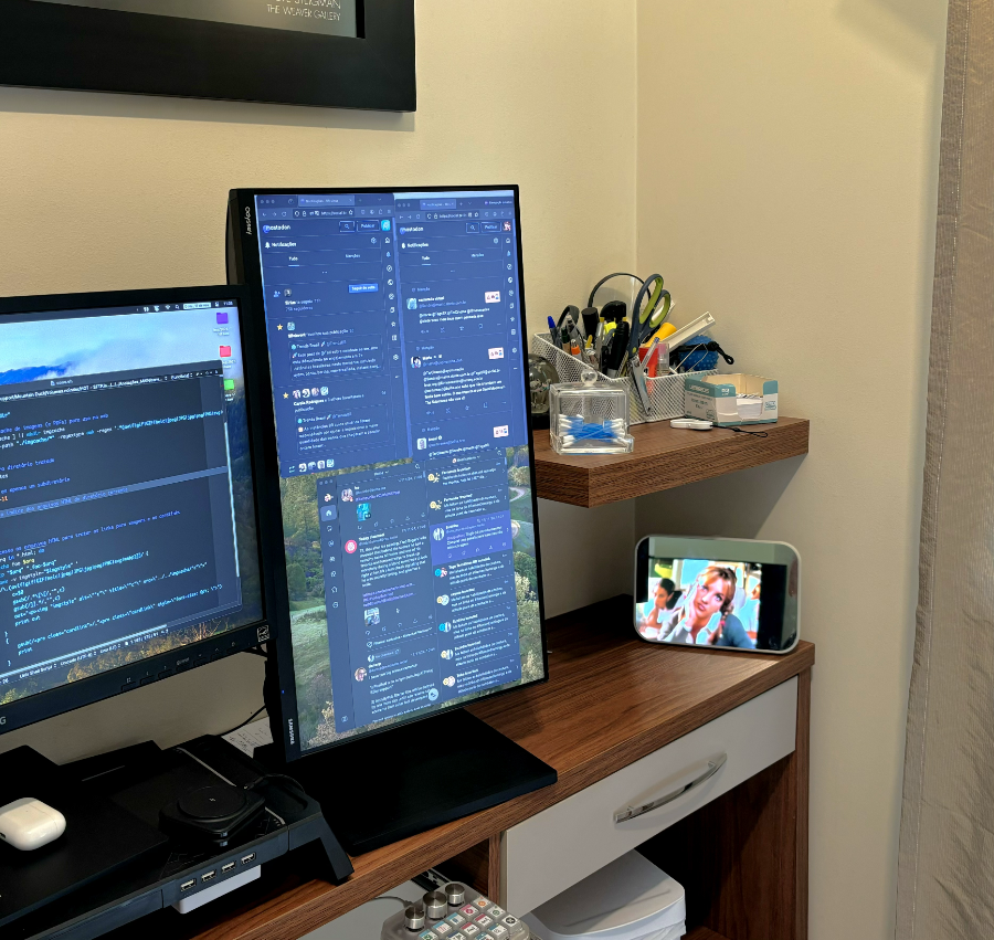 Visão parcial de uma mesa de trabalho, mostrando 2 monitores de computador e, um pouco afastado na lateral, um espqlho pequeno onde se pode ver a reflexão de uma tela de TV com um clip da Britney Spears no início da sua carreira.