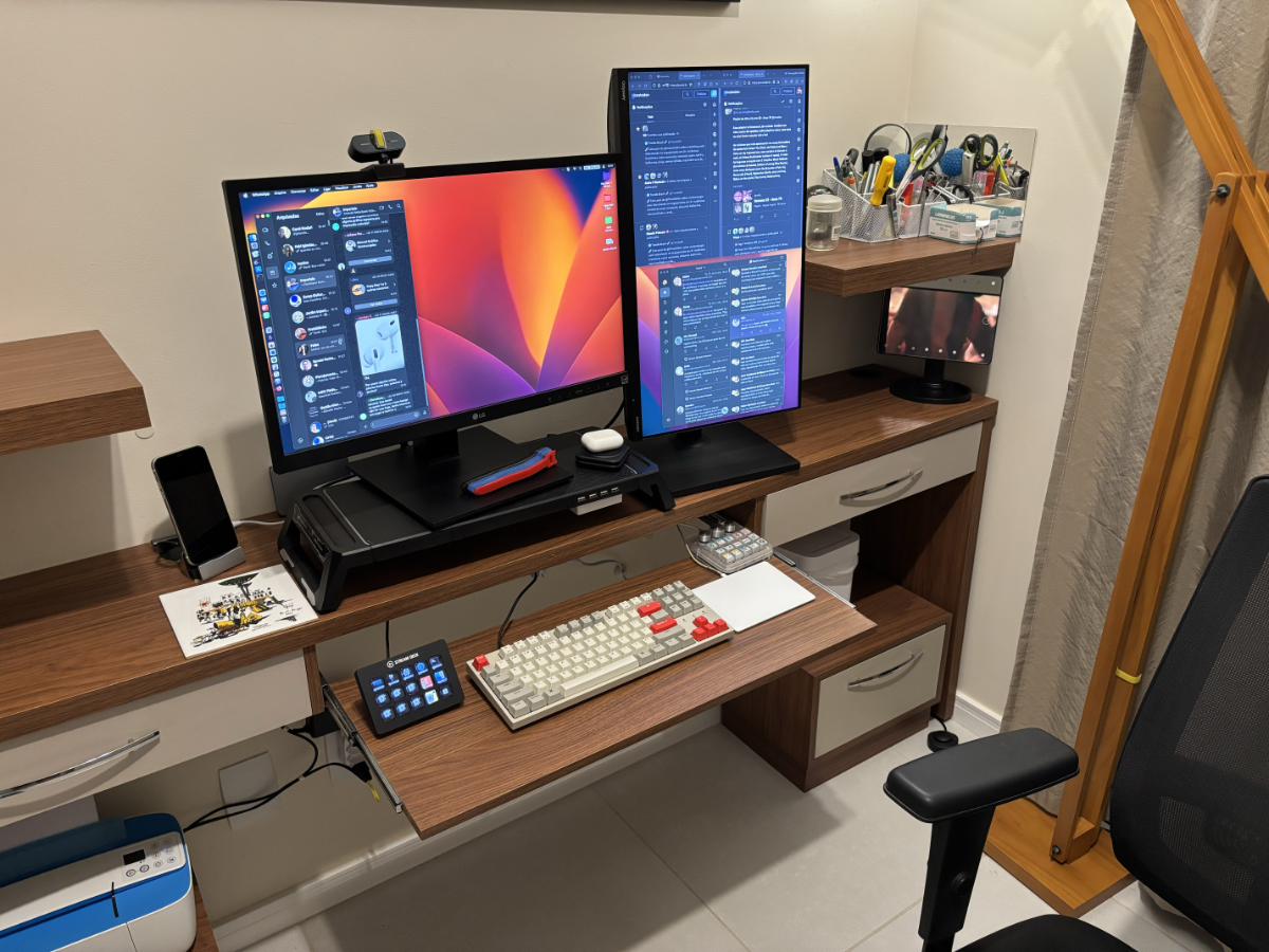 Foto da minha mesa de trabalho com 2 monitores, sendo um em orientação paisagem (mostrando uma janela do Whatsapp)  e outro em orientação retrato (mostrando várias janelas do Mastodon). <br><br>À frente deles estão 3 teclados e um trackpad, e ao lado está um espelho no qual aparece refletida uma cena de um clip da Rihanna.<br>