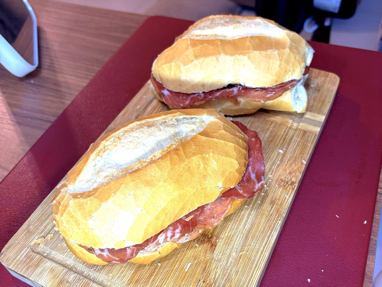 Dois sanduíches em cima de uma tábua. Os sanduíches são de pão francês, e é possível ver fatias finas de salame e de copa.