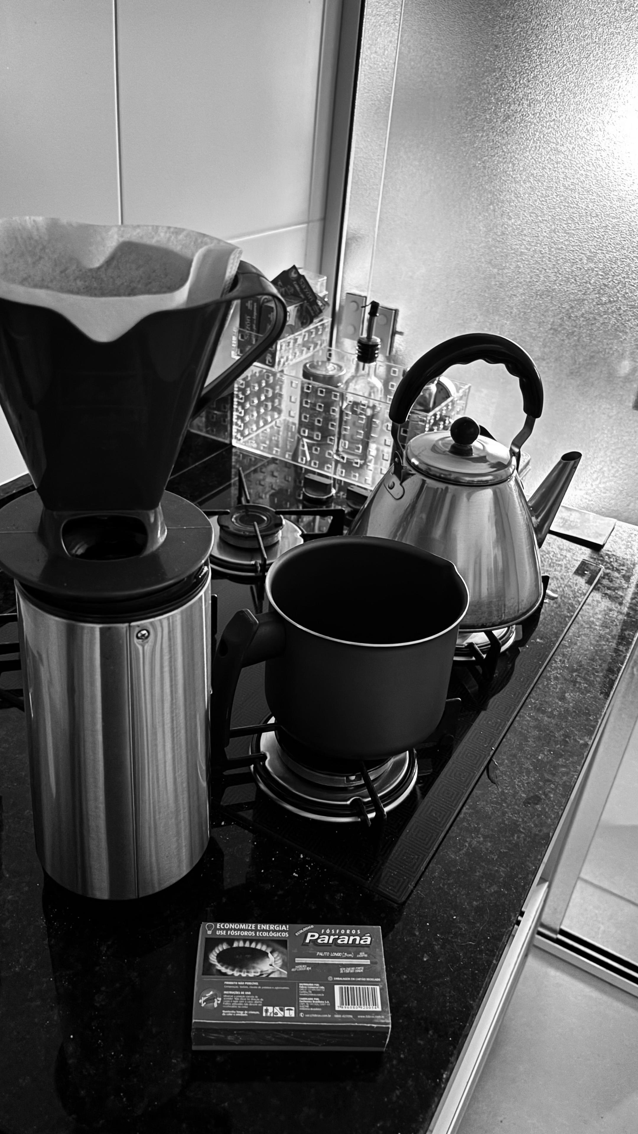 Foto em preto e branco mostrando um porta filtro posicionado sobre uma garrafa térmica e, ao fundo e sobre o fogão, uma chaleira e uma leiteira 
