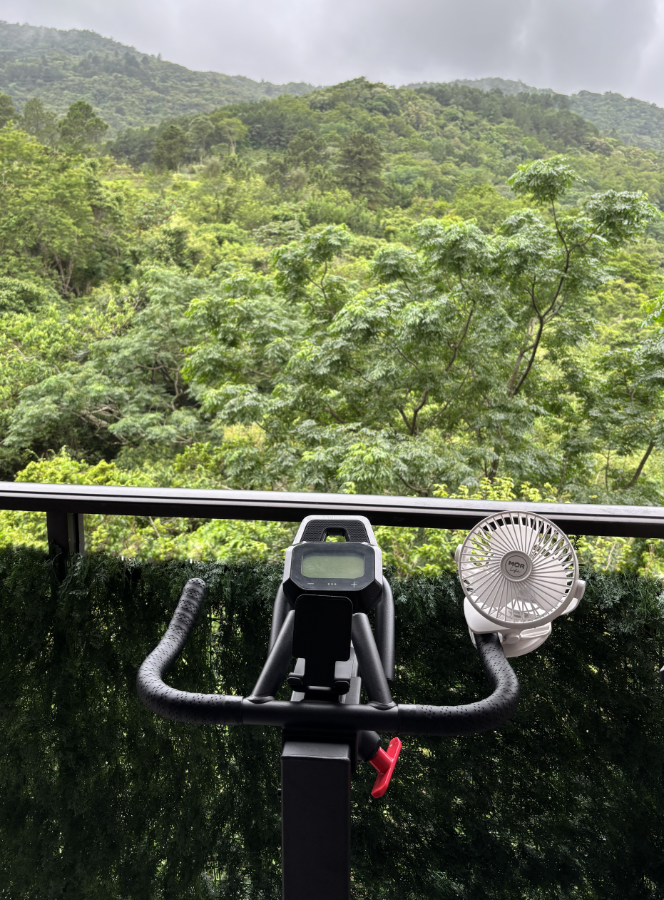 Foto do apoio de braço da bicicleta de spinning, ao qual foi fixado um ventilador portátil. Ele está posicionado junto ao guarda-corpo da sacada, e de frente para a mata e o morro.