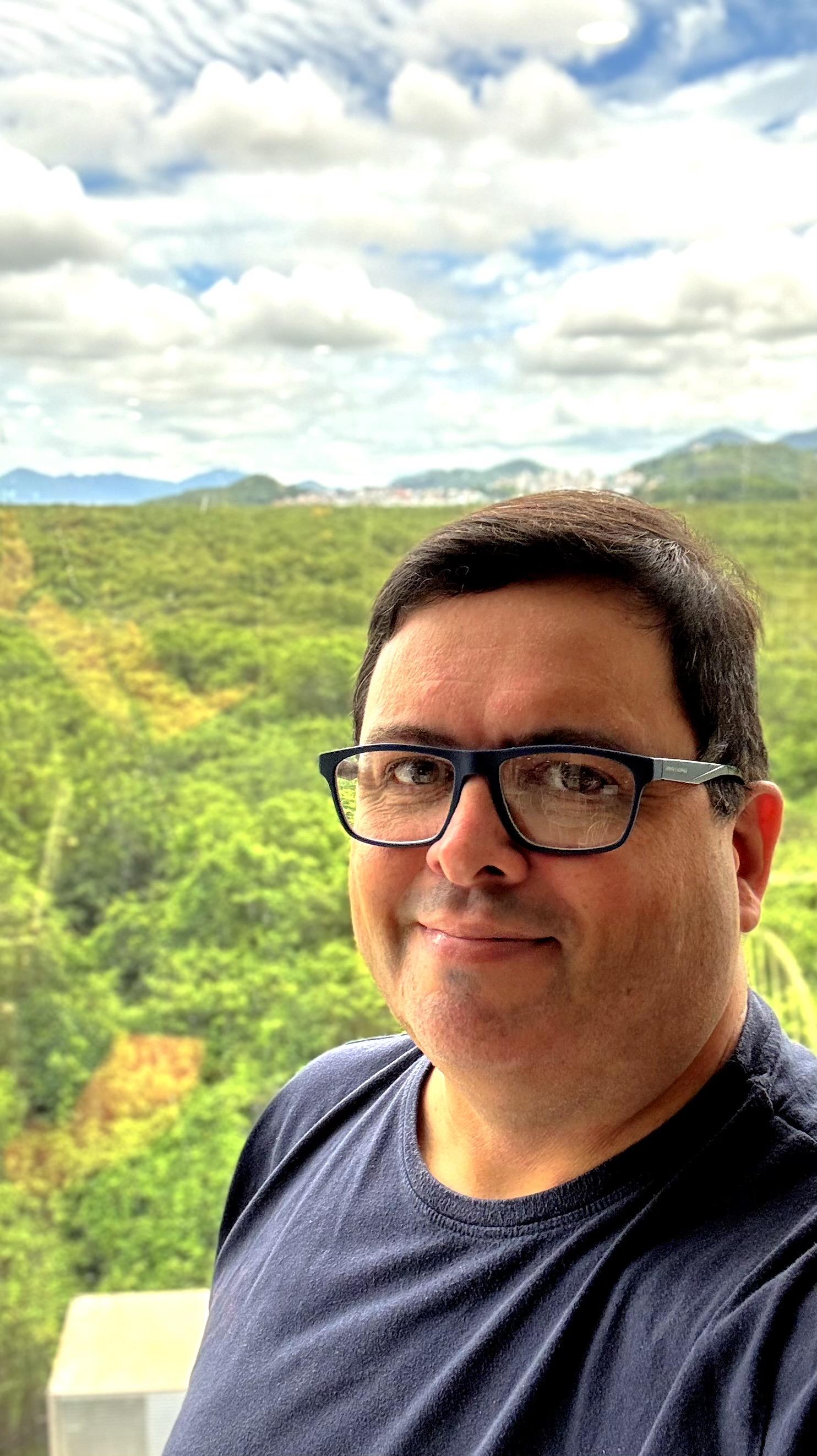 Foto minha, de camiseta azul, tendo ao fundo o manguezal do Itacorubi/Santa Mônica, e o céu nublado. 