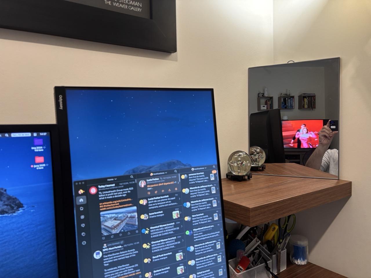 Foto mostrando um fragmento da minha mesa de trabalho, com o canto e a parte de cima de 2 monitores e, ao lado deles, um espelho posicionado de forma a exibir a TV que fica em outro ambiente (mostrando o clip de Material Girl, da Madonna).