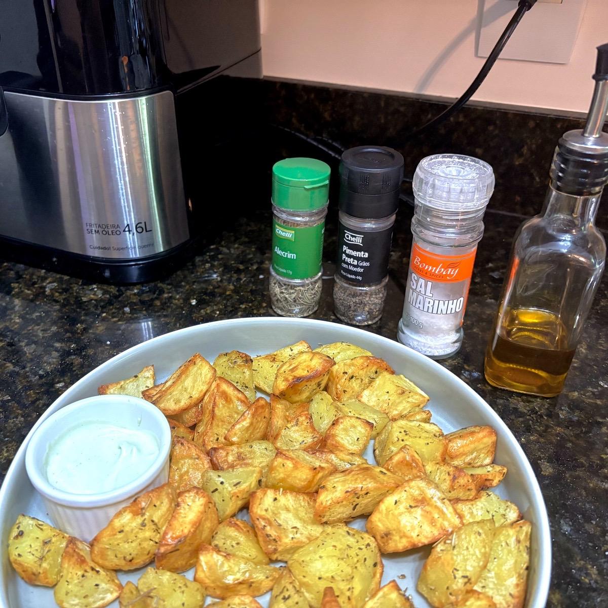 Travessa com batatas coradas e douradas, acompanhadas de uma cumbuquinha de maionese verde, e tendo ao fundo um vidro de azeite e os temperos a seguir: sal, pimenta do reino e alecrim, ao lado de uma air fryer.