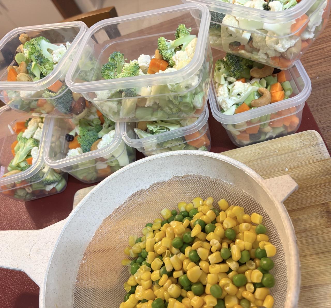 Em primeiro plano aparece uma peneira de cozinha, com uma quantidade de grãos de milho e de ervilha. Atrás, 7 potes transparentes, ainda sem tampa, cada um com uma mistura de cenoura, vagem, brócolis, couve-flor e castanhas.