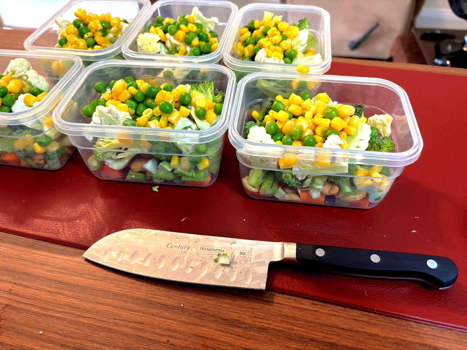 Uma faca de cozinha novinha em folha, mas já com sinais de ter sido usada recentemente, em frente a 6 potes herméticos, ainda abertos, cada um contendo um mix de cenoura, vagem, brócolis, couve flor, milho, ervilha e castanhas