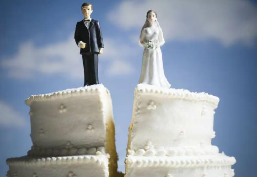 Um bolo no formato tradicional de bolo de casamento, rachado no meio e com os bonecos representando a noiva e o noivo posicionados em lados opostos da rachadura