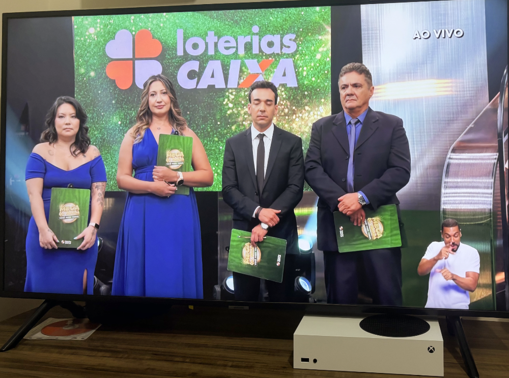 Imagem da transmissão do sorteio da Mega da Virada, mostrando os 4 auditores da Caixa e do Ministério da Fazenda, perfilados, incluindo a Maria Linux, à direita, de vestido azul, olhando séria para a câmera e segurando uma pasta.