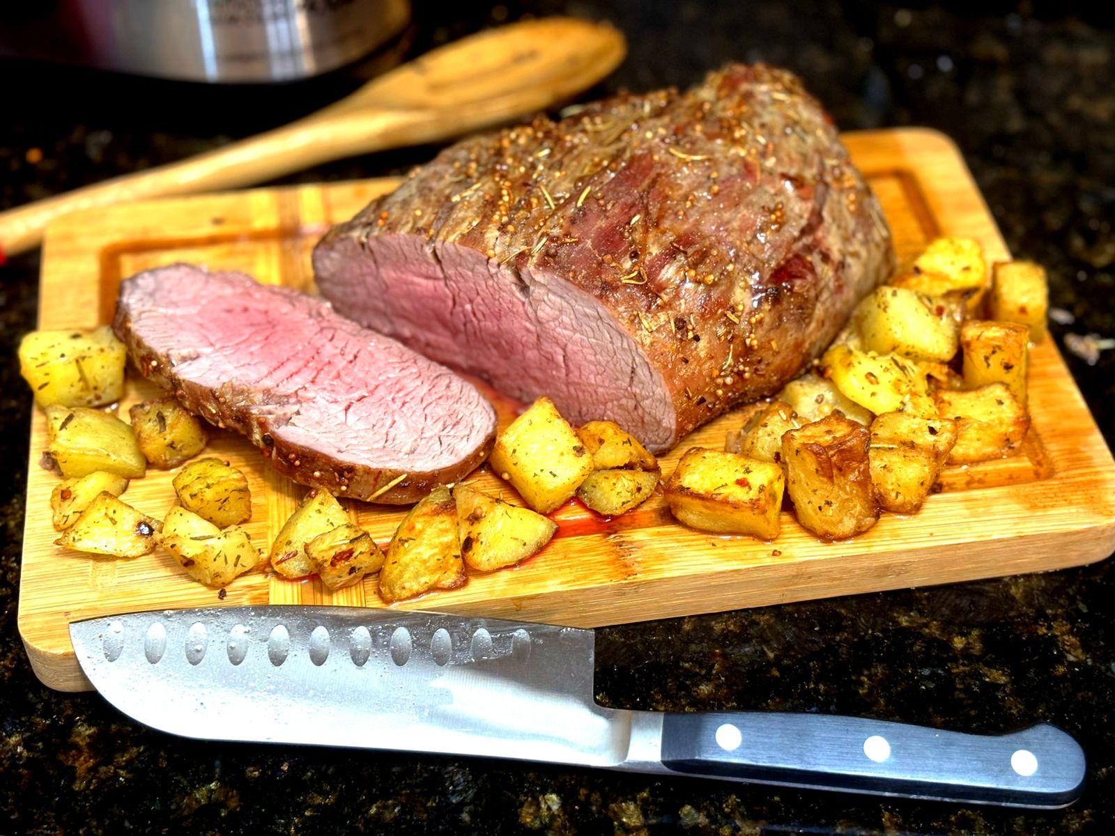 Foto de uma tábua de corte, sobre a qual está um rosbife pronto, ao ponto pra menos, com uma fatia já cortada, guarnecido com batatinhas.