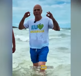 Frame do vídeo do prefeito, falando para a câmera enquanto caminha dentro da água, com uma camiseta branca com emblema nas cores verde, amarelo e azul, com o nome do município escrito em amarelo.