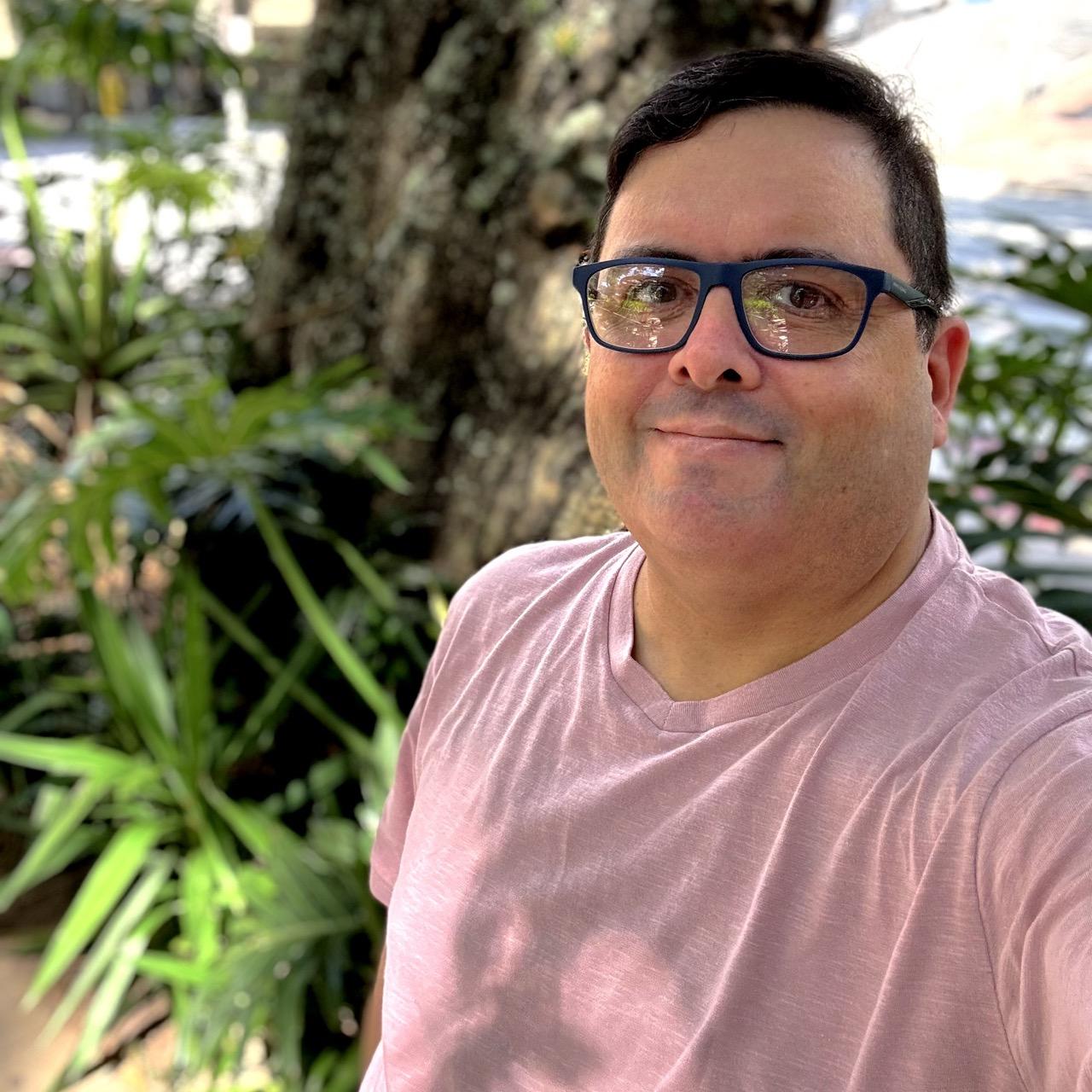 Foto minha, de camisa rosa-antigo e óculos com armação azul, olhando para a câmera, com a vegetação ao fundo.