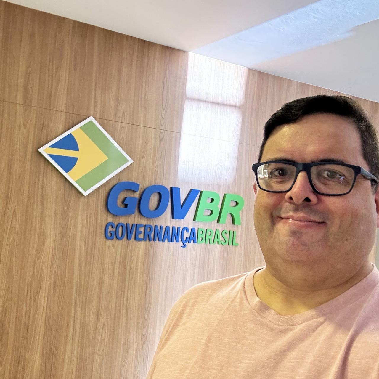 Foto minha olhando para a câmera, em frente a uma parede com o logo do govbr e as palavras Governança Brasil