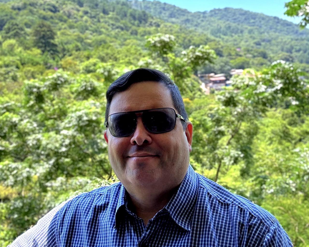 Foro minha, de camisa azul e óculos escuros, na sombra, tendo ao fundo a mata ensolarada