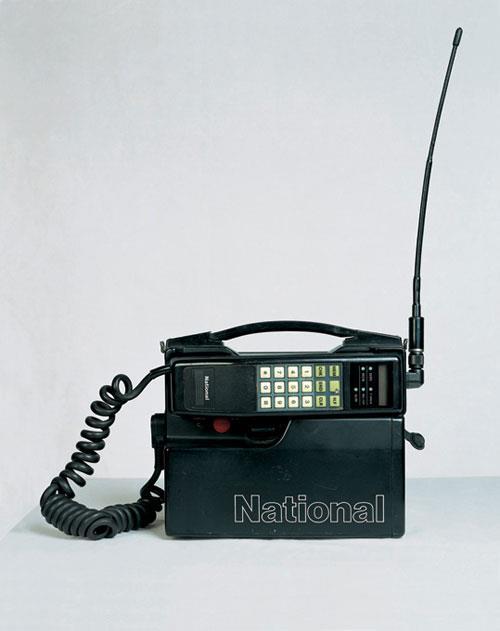 Foto de um dos primeiros telefones móveis com tecnologia similar à dos celulares de hoje - da National, com antena de 30 cm, e acoplado a uma bateria externa do tamanho de um paralelepípedo.