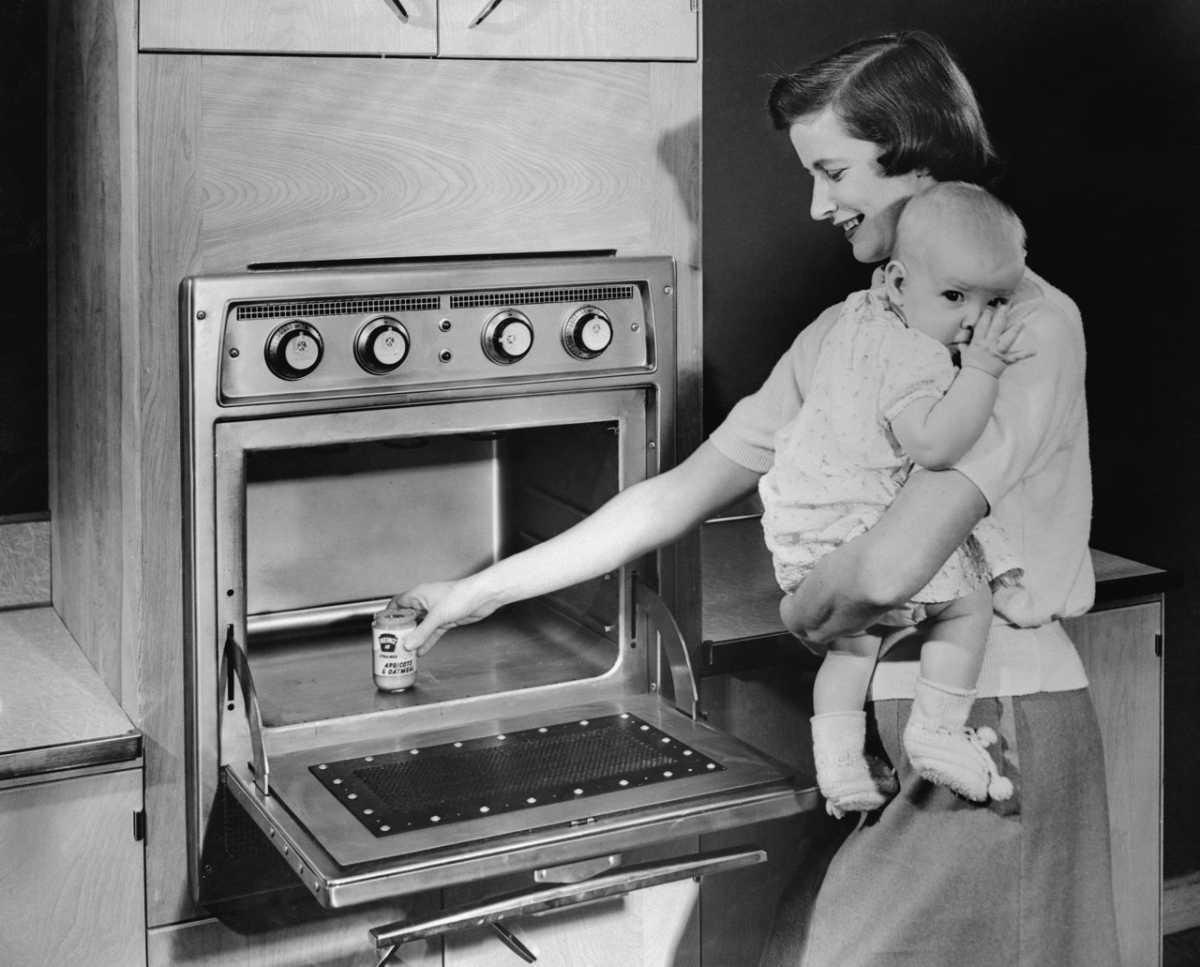 Um bebê no colo de sua mãe olha para a câmera, enquanto ela coloca um pote de papinha num gigantesco forno de microondas da década de 1950, em foto promocional em preto e branco.