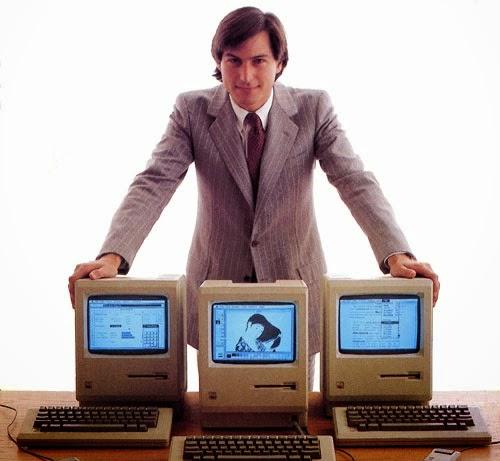 Steve Jobs, de terno risca de giz, junto a 3 Macs clássicos, em foto comercial.