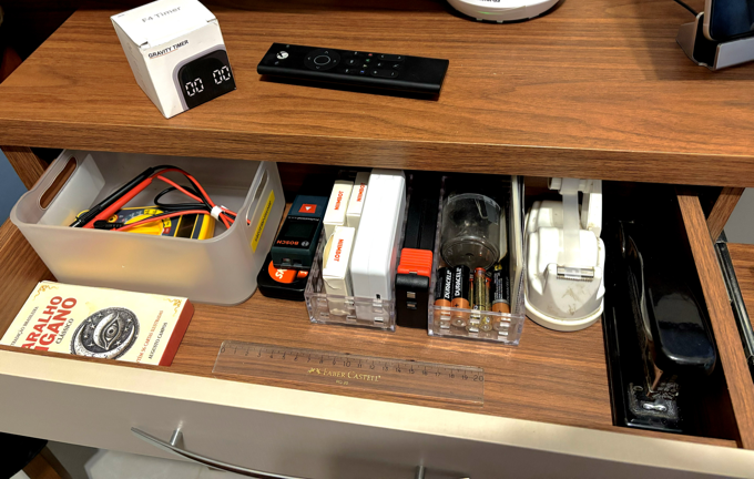 Foto de uma gaveta aberta e organizada, contendo, em sentido horário: um multímetro amarelo marca Hikari, duas trenas (uma digital e uma de fita), um rotulador niimbot com 3 caixas de fitas, um testador de pilhas, várias pilhas AA, rolo de fita adesiva, grampeador, régua e uma caixa de baralho cigano.