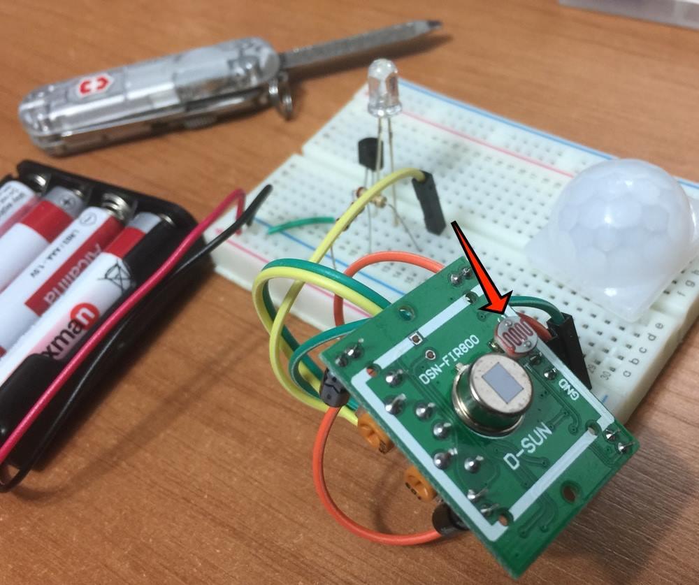 Foto de um sensor de presença simples, categoria hobby, conectado por vários fios a uma protoboard. O sensor foi modificado com a inserção de um componente, e uma seta sobreposta à foto indica a posição desse novo componente.