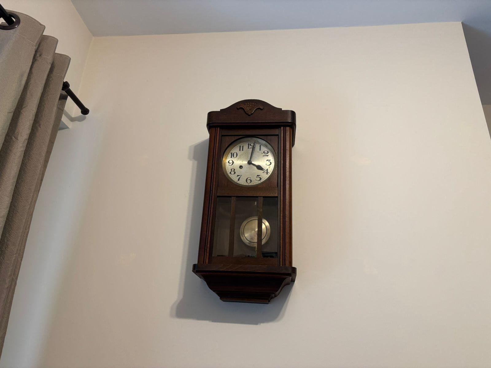 Um relógio de parede, em caixa de madeira decorada com um discreto entalhe em madeira, com os ponteiros marcando alguns minutos após as 16h.