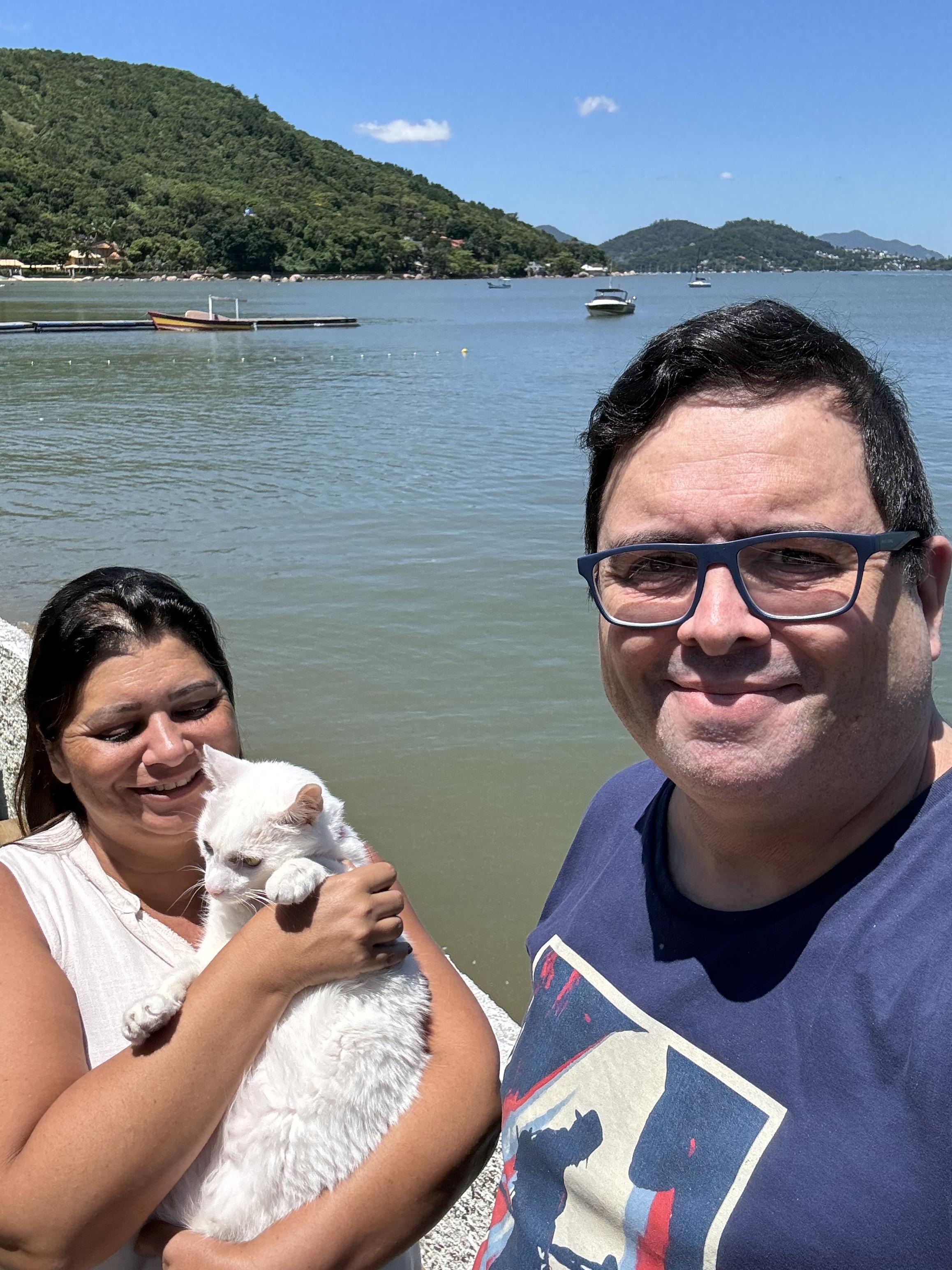 Com o mar ao fundo, uma mulher segura no colo um gato branco, ao lado de um homem de oculos e camiseta azul