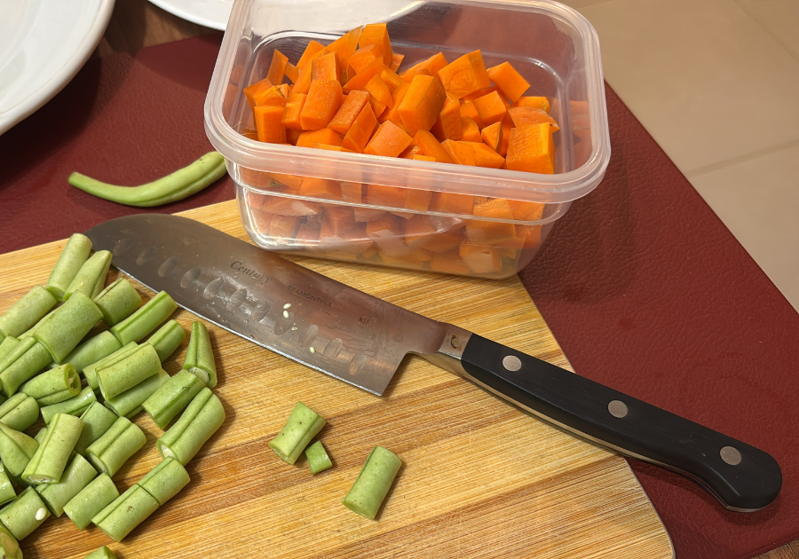 Uma faca sobre uma tábua de corte, entre algumas vagens já picadas, e um pote semitransparente contendo cenouras também picadas