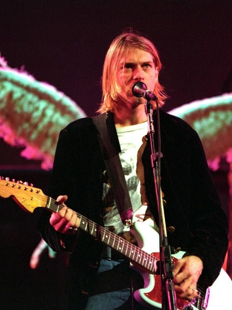 Kurt Cobain no palco, guitarra em mãos, posicionado de forma a interagir visualmente com a figura de um par de asas brancas do cenário.