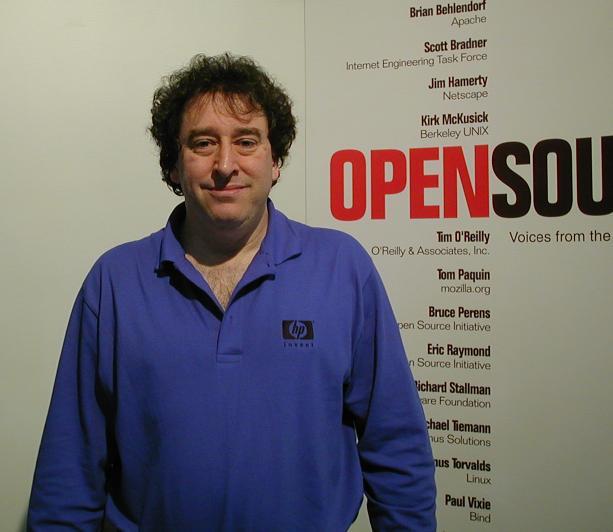 Foto de Bruce Perens, olhando para a câmera em frente a um poster em que está em destaque a expressão “Open Source“.